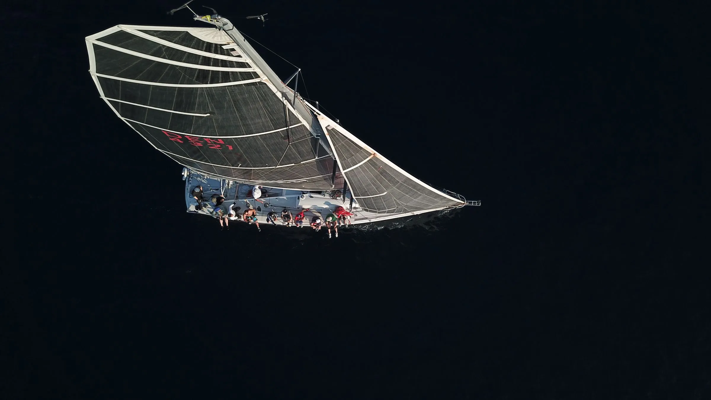 A sailboat with guests on deck, seen from a drone straight above dark water