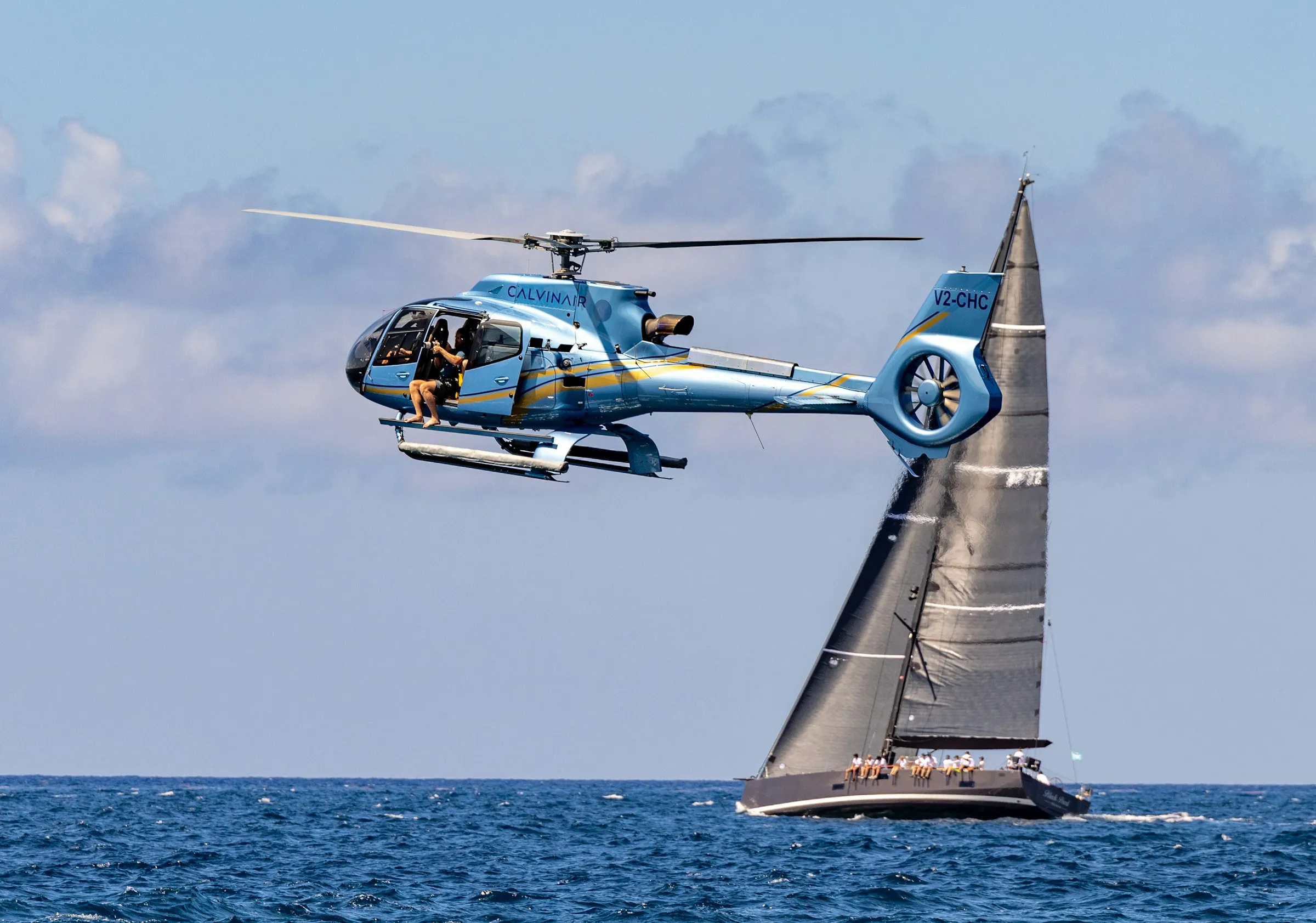A tour helicopter flying low over the ocean past a racing sailboat