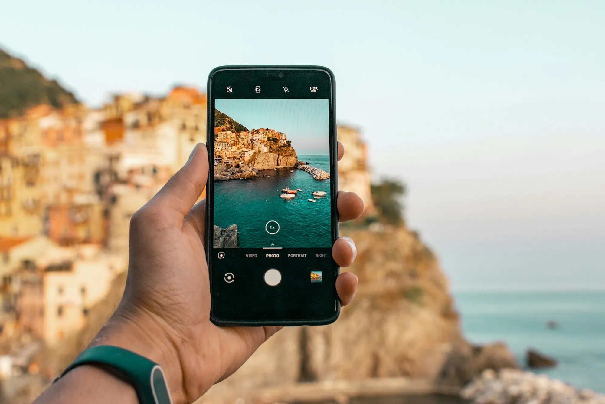 A hand holding a phone, taking a photo of a cliffside coastal village
