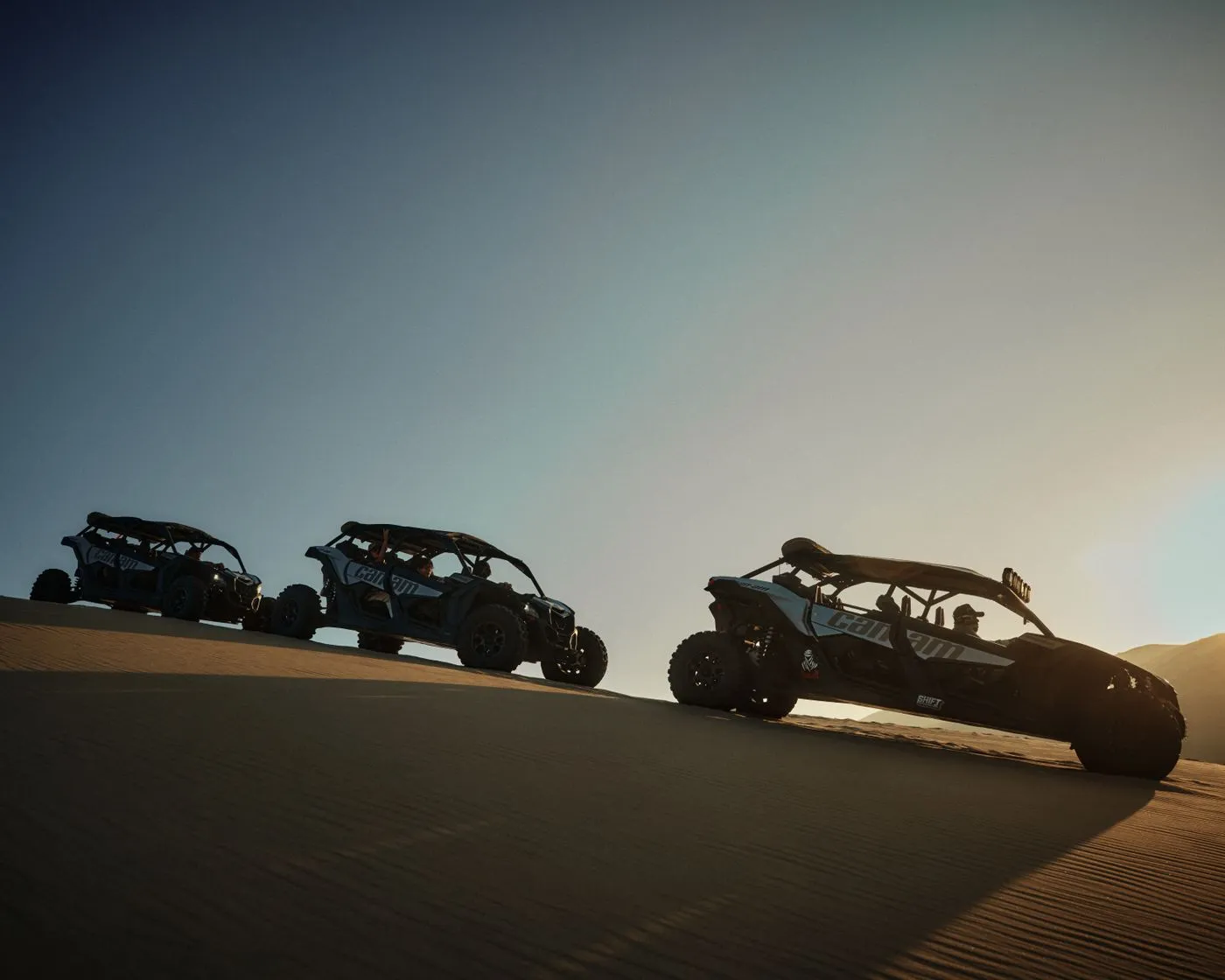 Three side-by-side off-road vehicles on a dune ridge at dusk